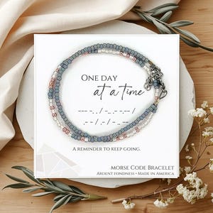 May include: A beaded bracelet with blue, white, and metallic beads on a white display card. The card features the text "ONE DAY AT A TIME," Morse code dots and dashes, "A REMINDER TO KEEP GOING," and "MORSE CODE BRACELET ARDENT FONDNESS MADE IN AMERICA."
