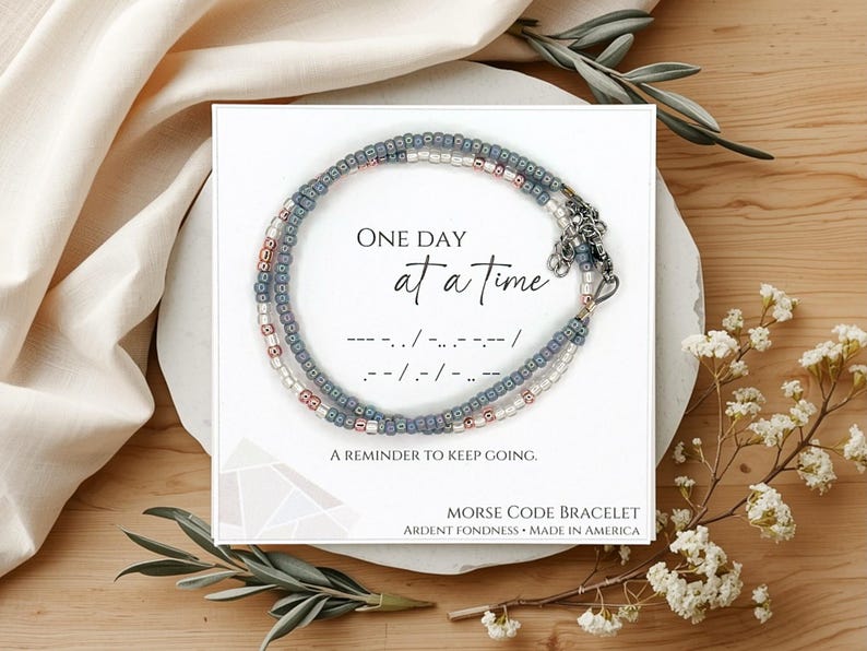 May include: A beaded bracelet with blue, white, and metallic beads on a white display card. The card features the text "ONE DAY AT A TIME," Morse code dots and dashes, "A REMINDER TO KEEP GOING," and "MORSE CODE BRACELET ARDENT FONDNESS MADE IN AMERICA."