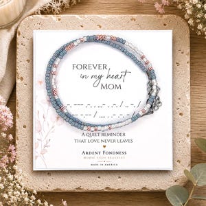 May include: A beaded bracelet featuring alternating blue, silver, and rose gold beads on a display card. The card reads: FOREVER in my heart MOM, includes Morse code, and the text: A QUIET REMINDER THAT LOVE NEVER LEAVES, ARDENT FONDNESS.