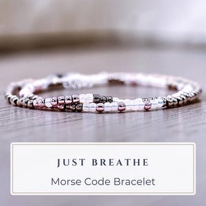 May include: A delicate Morse code bracelet with a wrap-around design. The bracelet features small, silver-colored beads interspersed with pink and dark beads. The text "JUST BREATHE" and "Morse Code Bracelet" are displayed in a white rectangle.