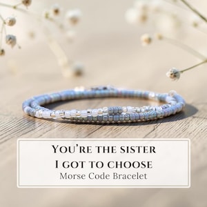 May include: A delicate beaded bracelet featuring a pattern of small, round, light blue, grey, and white beads. The text "YOU'RE THE SISTER I GOT TO CHOOSE Morse Code Bracelet" is printed in black on a white rectangular label positioned below the jewellery.