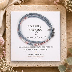 May include: A beaded bracelet featuring grey, white, and metallic pink seed beads arranged in a Morse code pattern. The bracelet is displayed on a white card with the text "YOU ARE enough" and "A REMINDER ON THE DAYS YOU FORGET. YOU ARE ALREADY ENOUGH. ARDENT FONDNESS MORSE CODE BRACELET MADE IN AMERICA."