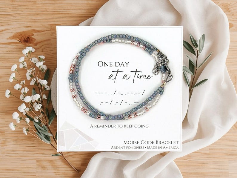 May include: A beaded bracelet featuring blue and metallic silver beads arranged in a circular pattern on a white display card. The card displays the text ONE DAY AT A TIME, Morse code symbols, A REMINDER TO KEEP GOING, and MORSE CODE BRACELET.