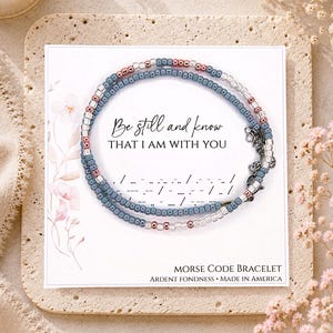 May include: A beaded bracelet featuring blue, silver, and copper-colored beads arranged in a Morse code pattern. The bracelet is displayed on a white card printed with the text Be still and know THAT I AM WITH YOU and MORSE CODE BRACELET.