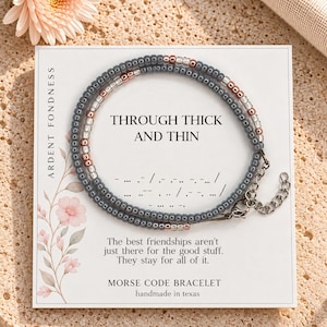 May include: A beaded bracelet featuring grey, clear, and metallic rose gold beads arranged in a pattern. The bracelet is displayed on a white card with the text "THROUGH THICK AND THIN," Morse code dots and dashes, and additional printed text.