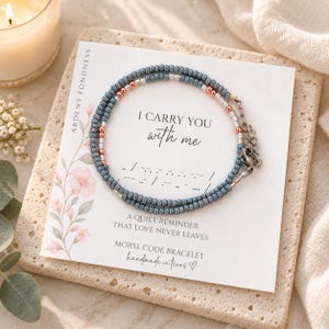 May include: A beaded bracelet with blue, silver, and copper-colored beads on a white display card. The card features the text I CARRY YOU WITH ME, Morse code dots and dashes, and the phrase A QUIET REMINDER THAT LOVE NEVER LEAVES.