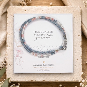 May include: A beaded bracelet featuring alternating blue, metallic rose gold, and clear glass beads on a display card. The card displays the text I HAVE CALLED YOU BY NAME, you are mine, Isaiah 43:1, and ARDENT FONDNESS MORSE CODE BRACELET.