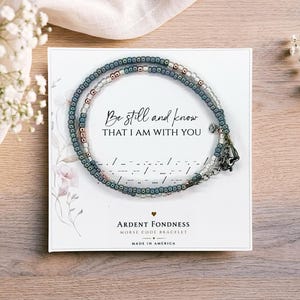 K&ouml;nnte beinhalten: Ein Perlenarmband mit blauen und metallischen Perlen, die kreisf&ouml;rmig auf einer wei&szlig;en Karte angeordnet sind. Die Karte zeigt den Text Be still and know THAT I AM WITH YOU, Morsecode-Symbole und den Markennamen Ardent Fondness.