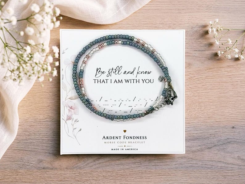 K&ouml;nnte beinhalten: Ein Perlenarmband mit blauen und metallischen Perlen, die kreisf&ouml;rmig auf einer wei&szlig;en Karte angeordnet sind. Die Karte zeigt den Text Be still and know THAT I AM WITH YOU, Morsecode-Symbole und den Markennamen Ardent Fondness.