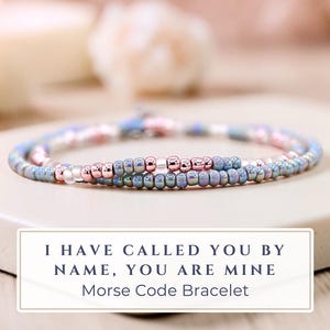 May include: A delicate beaded bracelet in shades of blue, pink, and iridescent beads. The bracelet is displayed on a white surface, with a sign that reads "I HAVE CALLED YOU BY NAME, YOU ARE MINE Morse Code Bracelet."
