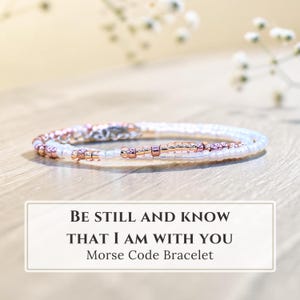 May include: A delicate bracelet featuring a pattern of small white and metallic rose gold beads arranged in a sequence. A rectangular label below the jewelry displays the text: BE STILL AND KNOW THAT I AM WITH YOU Morse Code Bracelet.
