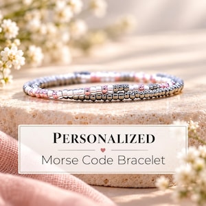May include: A delicate Morse code bracelet featuring a sequence of small metallic silver, dark grey, and soft pink beads. The bracelet is displayed with a rectangular sign that reads: PERSONALIZED Morse Code Bracelet with a small heart icon.
