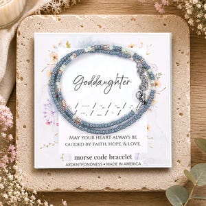 May include: A beaded bracelet featuring blue, white, and metallic rose gold seed beads arranged in a Morse code pattern. The bracelet is displayed on a white card printed with the word Goddaughter and the phrase May your heart always be guided by faith, hope, & love.