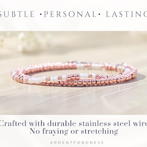K&ouml;nnte beinhalten: Ein Perlenarmband mit metallisch ros&eacute;goldenen und undurchsichtigen wei&szlig;en Saatperlen in einem sich wiederholenden Muster. Der Text SUBTLE &bull; PERSONAL &bull; LASTING steht dar&uuml;ber, und Crafted with durable stainless steel wire No fraying or stretching ARDENTFONDNESS darunter.