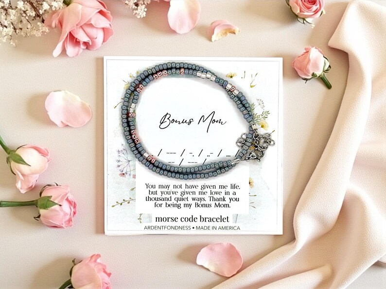 May include: A gray beaded Morse code bracelet with a silver clasp is displayed on a white card with the words "Bonus Mom." The card is surrounded by pink roses and petals. The bracelet is a thoughtful gift.