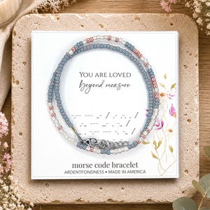 May include: A beaded bracelet featuring small blue, clear, and metallic rose gold beads arranged in a circular pattern on a white display card. The card displays the text YOU ARE LOVED beyond measure, Morse code dots and dashes, and morse code bracelet.