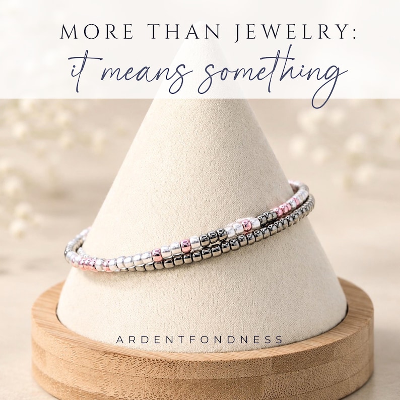 May include: A double-strand beaded bracelet featuring metallic silver, dark grey, and soft pink beads. The bracelet is displayed on a beige cone-shaped stand. Text at the top reads MORE THAN JEWELRY: it means something. Text at the bottom reads ARDENTFONDNESS.