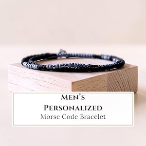 May include: A bracelet featuring a series of small, matte black and grey beads arranged in a pattern. The bracelet is displayed on a light-colored wooden block. The text on the image reads: MEN'S PERSONALIZED Morse Code Bracelet.