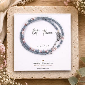 May include: A beaded bracelet featuring grey and metallic pink beads arranged in a Morse code pattern. The bracelet is displayed on a white card with the text "let them" and "ARDENT FONDNESS MORSE CODE BRACELET MADE IN AMERICA" printed on it.