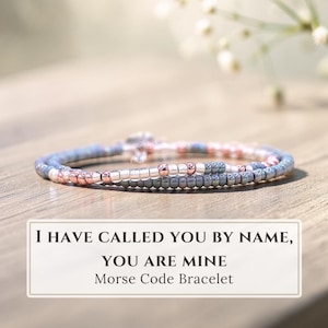 Peut inclure: Un bracelet composé de petites perles grises, rose gold métallisées et blanches disposées en séquence. Le texte I HAVE CALLED YOU BY NAME, YOU ARE MINE Morse Code Bracelet est affiché dans un cadre rectangulaire sous l'objet présenté.