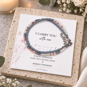 May include: A delicate beaded bracelet featuring alternating blue, silver, and rose gold beads arranged in a Morse code pattern. The bracelet is displayed on a white card printed with the text I CARRY YOU with me, A QUIET REMINDER THAT LOVE NEVER LEAVES, and ARDENT FONDNESS MORSE CODE BRACELET.
