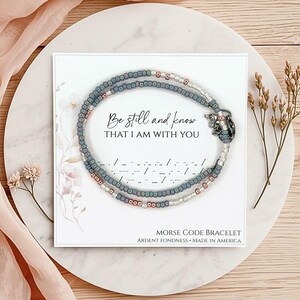 K&ouml;nnte beinhalten: Ein Perlenarmband mit grauen, metallischen und wei&szlig;en Saatperlen, die kreisf&ouml;rmig auf einer quadratischen Karte angeordnet sind. Die Karte zeigt den Text Be still and know THAT I AM WITH YOU, Morsecode-Symbole und MORSE CODE BRACELET.