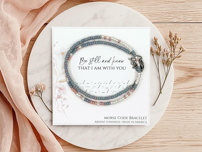 K&ouml;nnte beinhalten: Ein Perlenarmband mit grauen, metallischen und wei&szlig;en Saatperlen, die kreisf&ouml;rmig auf einer quadratischen Karte angeordnet sind. Die Karte zeigt den Text Be still and know THAT I AM WITH YOU, Morsecode-Symbole und MORSE CODE BRACELET.