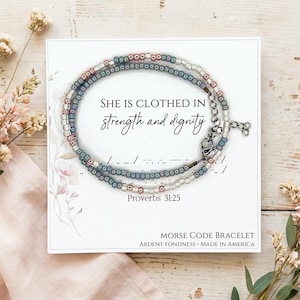 May include: A Morse code bracelet with a silver clasp and a mix of white, blue, and pink beads. The bracelet is displayed on a white card with the text "SHE IS CLOTHED IN strength and dignity" and "Proverbs 31:25".