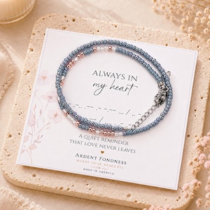 May include: A beaded bracelet featuring small blue and rose gold metallic beads arranged in a pattern, displayed on a white card with the text "ALWAYS IN MY HEART" and "A QUIET REMINDER THAT LOVE NEVER LEAVES" along with Morse code symbols.