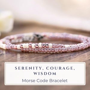 May include: A delicate beaded bracelet featuring a repeating pattern of metallic mauve and opaque white seed beads. The bracelet includes a rectangular white label with the text: SERENITY, COURAGE, WISDOM. MORSE CODE BRACELET.