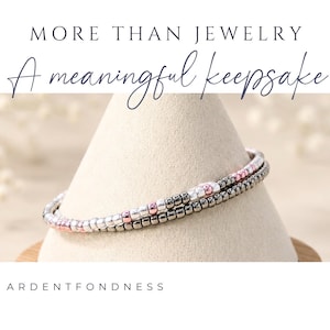 K&ouml;nnte beinhalten: Ein zweireihiges Perlenarmband mit kleinen metallischen silbernen, ros&eacute;goldenen und dunkelgrauen Perlen. Der Text MORE THAN JEWELRY A meaningful keepsake und ARDENTFONDNESS ist &uuml;ber und unter dem Armband auf dem Bild zu sehen.