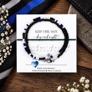 May include: A Morse code bracelet made of small black, white, and blue beads on a white display card. The card features the text KEEP HER SAFE day and night, Morse code symbols, a blue heart icon, and the text MORSE CODE BRACELET ARDENTFONDNESS MADE IN AMERICA.
