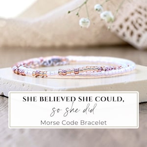 May include: A delicate beaded bracelet featuring a pattern of white, metallic rose gold, and soft pink seed beads. A rectangular white label is overlaid with the black text: SHE BELIEVED SHE COULD, so she did, Morse Code Bracelet.
