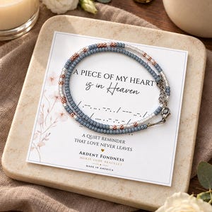 May include: A Morse code bracelet featuring small blue, white, and metallic copper beads on a display card. The card reads: A PIECE OF MY HEART is in Heaven, A QUIET REMINDER THAT LOVE NEVER LEAVES, ARDENT FONDNESS MORSE CODE BRACELET, MADE IN AMERICA.