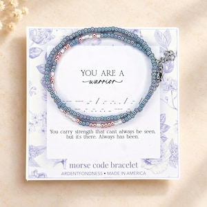 May include: A beaded bracelet featuring blue and metallic rose gold beads arranged in a Morse code pattern. The bracelet is displayed on a white card printed with the text YOU ARE A warrior and You carry strength that can't always be seen.