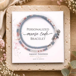 May include: A delicate bracelet made of small blue, pink, and clear beads arranged in a pattern, displayed on a white card with the text Personalized Morse Code Bracelet, Ardent Fondness, and Made in America printed in black and gold lettering.