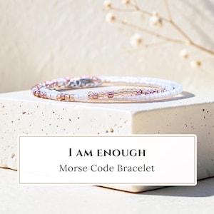 May include: A Morse code bracelet featuring a delicate arrangement of small white and metallic rose gold beads. The text I AM ENOUGH Morse Code Bracelet is displayed in a rectangular frame below the jewelry piece, which rests on a block.