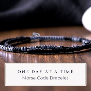 May include: A Morse code bracelet featuring a sequence of matte black and grey beads with metallic accents. The text "ONE DAY AT A TIME" and "Morse Code Bracelet" is printed in a white rectangular box at the bottom of the image.
