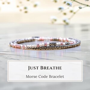 Peut inclure: Un bracelet délicat composé de perles métalliques couleur or rose, argent et gris foncé. Le texte "JUST BREATHE" et "Morse Code Bracelet" est imprimé dans un encadré rectangulaire situé sous le bijou sur l'image présentée.