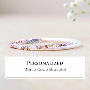 May include: A delicate bracelet featuring a sequence of small white and metallic rose gold beads arranged in a pattern. The text "PERSONALIZED Morse Code Bracelet" is displayed in a white rectangular box below the jewelry item.