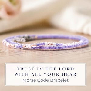 May include: A delicate, beaded bracelet in shades of lavender and white, with rose gold accents. The bracelet is coiled and rests on a light wooden surface. Text on the image reads: "Trust in the Lord with all your heart. Morse Code Bracelet."