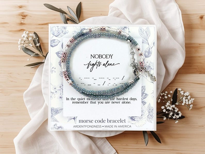 May include: A Morse code bracelet featuring small blue and metallic beads on a display card with the text NOBODY fights alone, In the quiet moments and the hardest days, remember that you are never alone, and morse code bracelet ARDENTFONDNESS.
