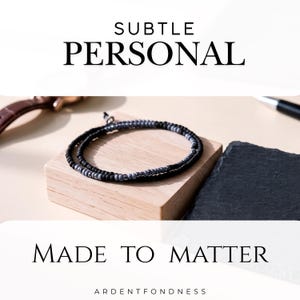 May include: A wrap bracelet featuring alternating matte black and metallic grey beads. The text SUBTLE PERSONAL is printed at the top, and MADE TO MATTER ARDENTFONDNESS is printed at the bottom. The bracelet is coiled in a circular shape.
