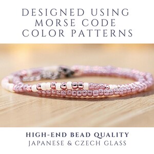 May include: A beaded bracelet featuring a repeating pattern of metallic rose gold and opaque white glass beads arranged in a Morse code sequence. The text above reads DESIGNED USING MORSE CODE COLOR PATTERNS and below HIGH-END BEAD QUALITY JAPANESE & CZECH GLASS.