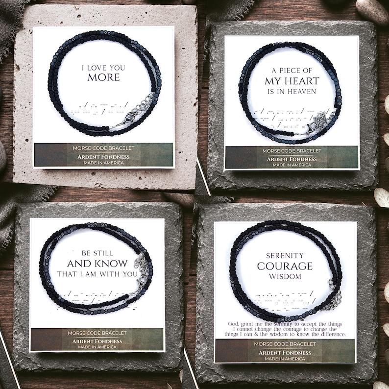 May include: Four black beaded Morse code bracelets displayed on white cards. Each card features a unique phrase: I LOVE YOU MORE, A PIECE OF MY HEART IS IN HEAVEN, BE STILL AND KNOW THAT I AM WITH YOU, and SERENITY COURAGE WISDOM. Each bracelet has a metal clasp.