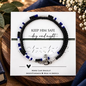 May include: A beaded bracelet featuring a pattern of black, white, and blue beads on a white display card. The card displays the text KEEP HIM SAFE day and night, Morse code symbols, a heart icon, and the text MORSE CODE BRACELET ARDENTFONDNESS MADE IN AMERICA.
