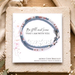 K&ouml;nnte beinhalten: Ein Morsecode-Armband mit blauen, silbernen und kupferfarbenen Perlen auf einer wei&szlig;en Pr&auml;sentationskarte. Die Karte zeigt den Text Be still and know THAT I AM WITH YOU, eine Morsecode-Sequenz und die Worte MORSE CODE BRACELET.