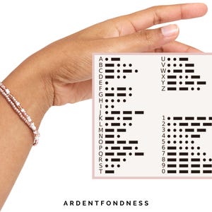 Peut inclure: Un bracelet &agrave; deux rangs avec des perles roses et blanches est port&eacute; au poignet. Une carte affiche le code Morse pour l'alphabet et les chiffres. Le texte "ARDENTFONDNESS" est en bas de l'image.