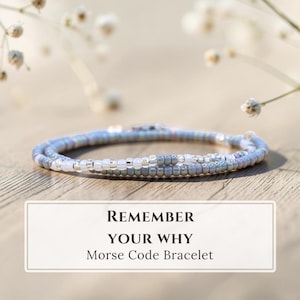 Peut inclure: Un bracelet délicat composé de perles de rocaille dans des tons bleu clair, gris et blanc disposées en motif. Une étiquette rectangulaire sous le bracelet affiche le texte : REMEMBER YOUR WHY Morse Code Bracelet en lettres noires.