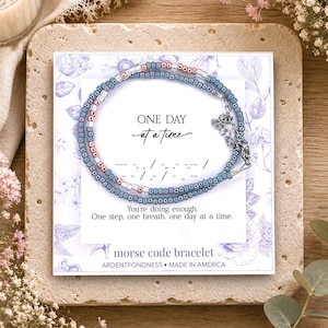May include: A Morse code bracelet featuring small blue and metallic rose gold beads on a display card. The card displays the text ONE DAY at a time, a Morse code sequence, and the phrase You're doing enough. One step, one breath, one day at a time.